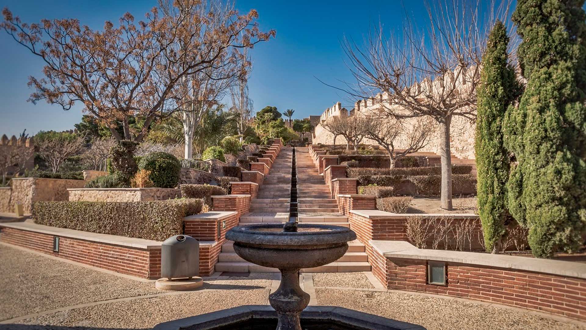 Una fuente con varios caminos hechos de ladrillos y árboles decorando los jardines de la Alcazaba